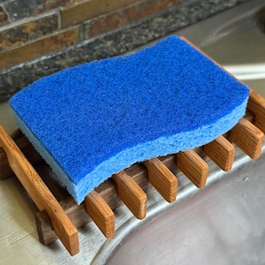 Puede incluir: Una esponja de cocina azul y blanca descansa sobre un escurridor de madera. El escurridor es de madera marrón claro y tiene un diseño de listones. La esponja es rectangular y parece nueva.