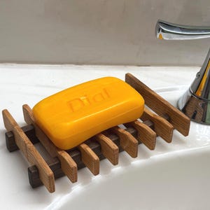 May include: A wooden soap dish with a dark brown base and light brown slats holds a yellow bar of Dial soap. The soap dish is placed on a white sink, near a chrome faucet.