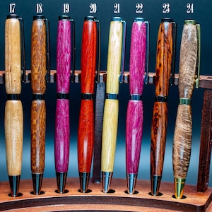 Puede incluir: Una colección de bolígrafos de madera hechos a mano en varios colores y vetas de madera, expuestos en un portalápices de madera. Los bolígrafos presentan detalles en negro y dorado, con una variedad de colores como marrón claro, marrón oscuro, rosa, rojo y amarillo.