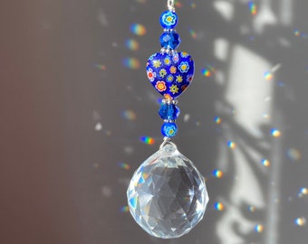 Op de afbeelding: Een kristallen zonenvanger met een heldere kristallen bol en een blauwe hartvormige hanger met een bloemenpatroon. De hartvormige hanger is bevestigd aan een zilveren ketting.