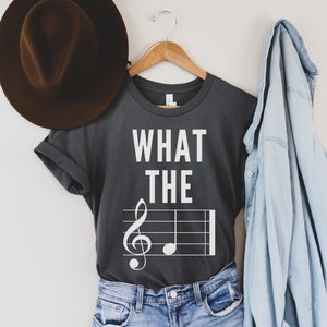 May include: A gray t-shirt with the text "WHAT THE" and a treble clef on a musical staff. The shirt is hanging on a wooden hanger with a brown hat and a light blue denim shirt on the side.