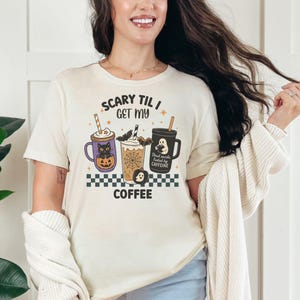 May include: Cream-colored t-shirt with the text "Scary Til I Get My Coffee" and a Halloween-themed graphic of three coffee cups. The cups feature a cat, a bat, and a ghost. A black and white checkered pattern is at the bottom.