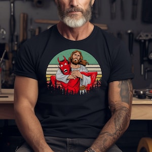 Könnte beinhalten: Schwarzes T-Shirt mit einer Grafik von Jesus, der einen roten Teufel umarmt. Das Design ist vor einem Retro-Sonnenuntergangshintergrund mit grünen, gelben und beigen Farben platziert. Eine Waldsilhouette befindet sich am unteren Rand.