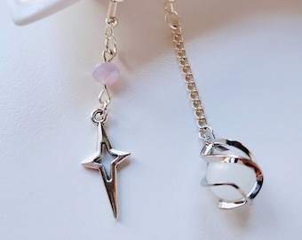 May include: Asymmetrical earrings. One earring has a silver star charm and a pink bead. The other features a white orb in a silver cage, suspended from a chain. The earrings are displayed on a white surface.