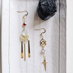 May include: A pair of gold earrings with dangling charms. One earring features a red gemstone, a filigree design, and three thin gold bars. The other earring has a geometric design, a clear rhinestone, and a star-shaped charm.