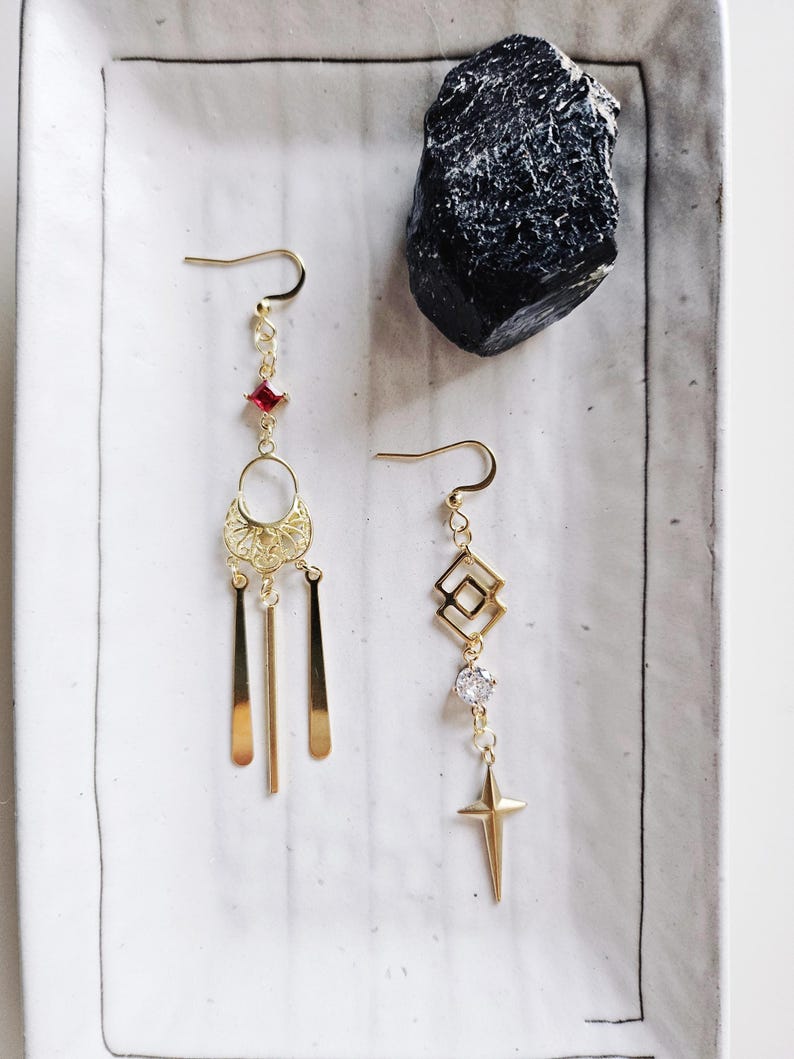 May include: A pair of gold earrings with dangling charms. One earring features a red gemstone, a filigree design, and three thin gold bars. The other earring has a geometric design, a clear rhinestone, and a star-shaped charm.