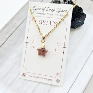 May include: A gold-colored chain necklace with a star-shaped pendant. The pendant is a reddish-brown color. The necklace is displayed on a white card with the text "SYLUS" and "Eyes of Deep Space".