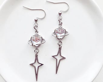 May include: A pair of silver-coloured drop earrings. Each earring has a heart-shaped charm with a planet-like ring, and a star-shaped pendant. The earrings are displayed on a white plate.