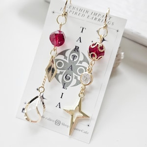 May include: Gold earrings inspired by the Genshin Impact video game. The earrings feature a red crystal, a gold star, a clear crystal, and a gold bead with a red design. The earrings are hanging from a gold hook.