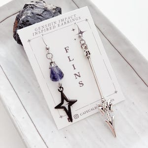 May include: A pair of Genshin Impact inspired earrings. One earring features a dark blue crystal and a black star. The other earring has a silver spearhead design. The earrings are displayed on a white card with text that reads "FLINS" and "GENSHIN IMPACT INSPIRED EARRINGS".