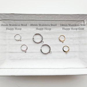 May include: A rectangular white ceramic tray displays several huggy hoop earrings. The earrings are silver and gold-colored, with sizes including 14mm and 18mm. Text on the tray indicates the material is stainless steel.