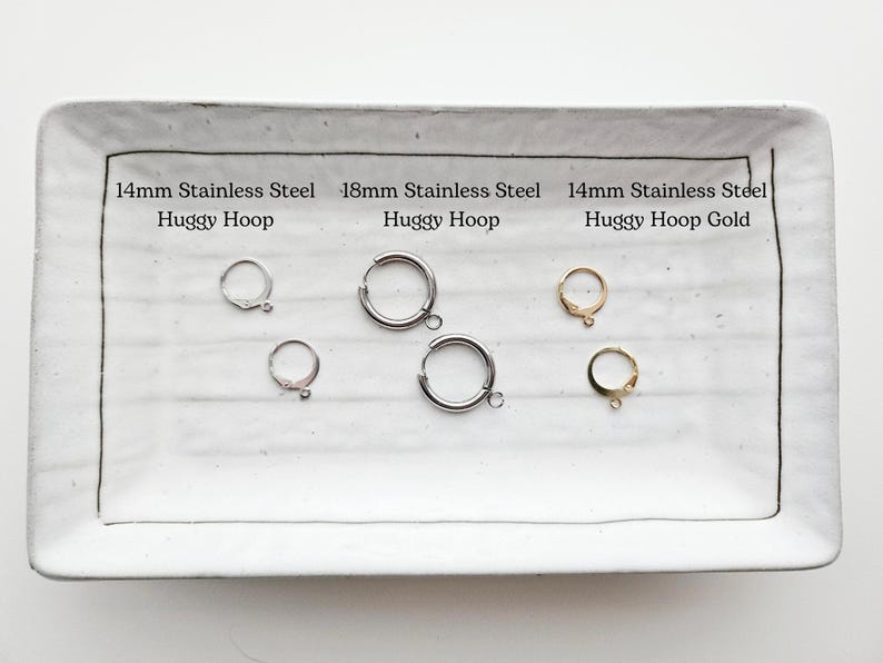 May include: A rectangular white ceramic tray displays several huggy hoop earrings. The earrings are silver and gold-colored, with sizes including 14mm and 18mm. Text on the tray indicates the material is stainless steel.