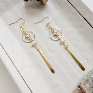 K&ouml;nnte beinhalten: Goldene Ohrh&auml;nger mit einem kleinen quadratischen Strassstein, einem Kreis und einem Stern-Anh&auml;nger. Die Ohrringe haben eine lange, d&uuml;nne, goldene Stange, die von unten h&auml;ngt.