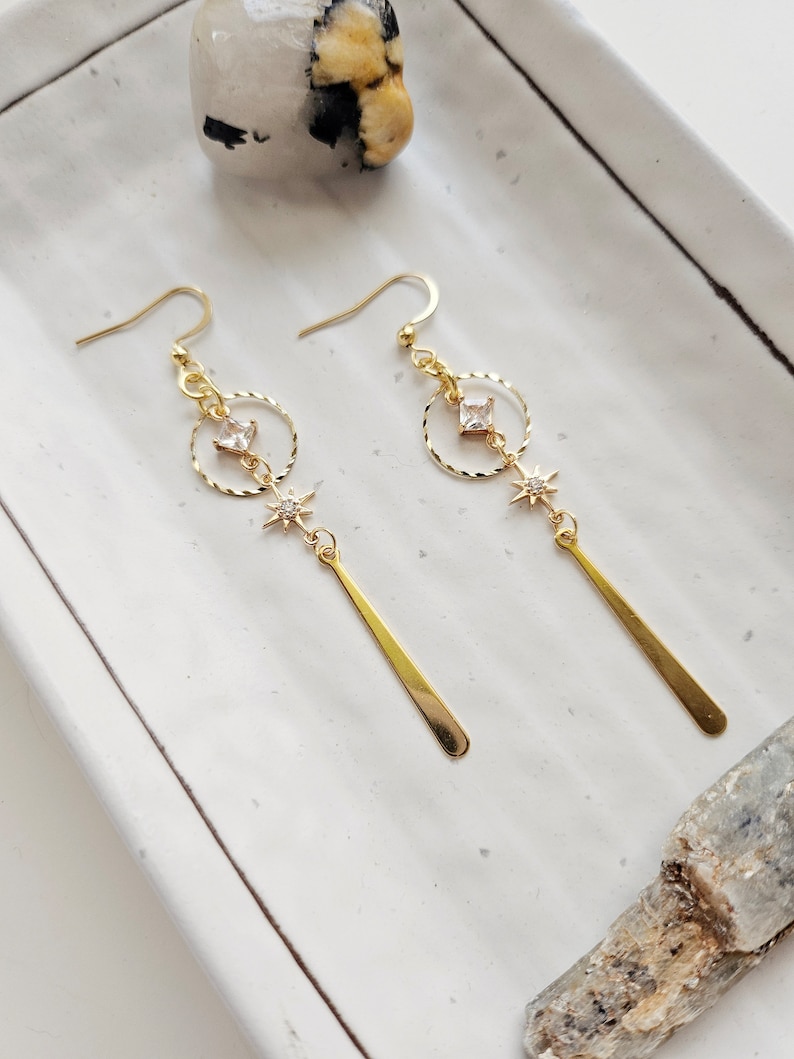 K&ouml;nnte beinhalten: Goldene Ohrh&auml;nger mit einem kleinen quadratischen Strassstein, einem Kreis und einem Stern-Anh&auml;nger. Die Ohrringe haben eine lange, d&uuml;nne, goldene Stange, die von unten h&auml;ngt.