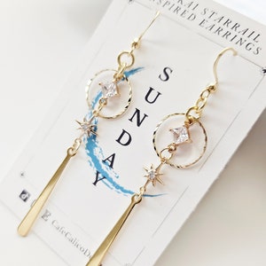 K&ouml;nnte beinhalten: Goldene Ohrringe mit einem Stern und einem quadratischen Strassstein-Anh&auml;nger, der an einem Kreis h&auml;ngt. Die Ohrringe sind mit "Honkai Starrail Inspired Earrings" und "Sunday" beschriftet.