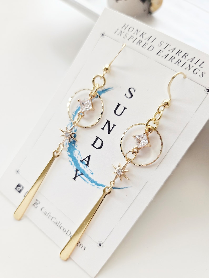 K&ouml;nnte beinhalten: Goldene Ohrringe mit einem Stern und einem quadratischen Strassstein-Anh&auml;nger, der an einem Kreis h&auml;ngt. Die Ohrringe sind mit "Honkai Starrail Inspired Earrings" und "Sunday" beschriftet.