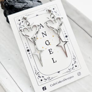 May include: Silver-toned earrings with heart and star designs. The earrings are displayed on a white card with the word "ANGEL" printed on it. The card also features the CafeCalicoDesigns logo.