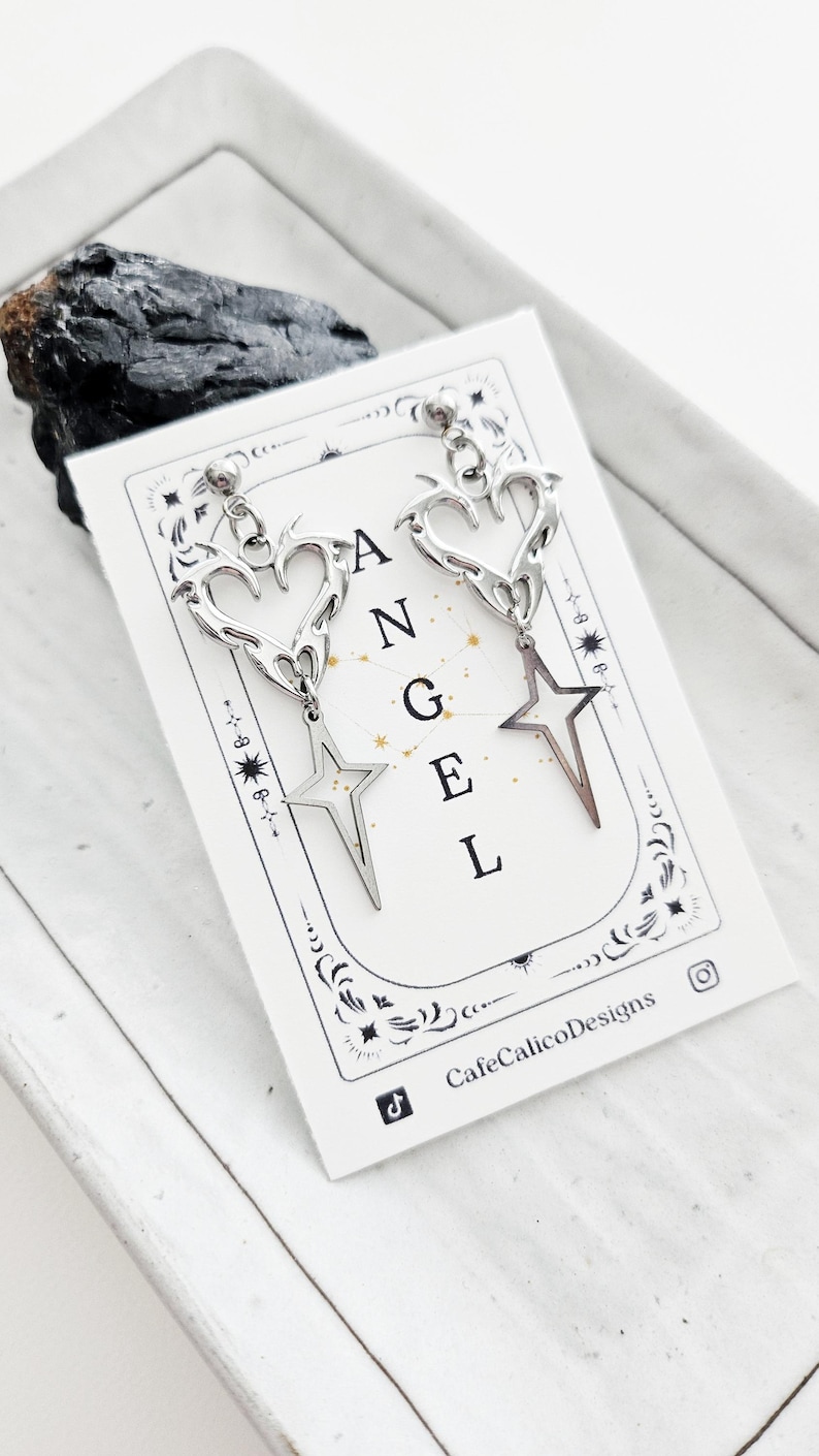 May include: Silver-toned earrings with heart and star designs. The earrings are displayed on a white card with the word "ANGEL" printed on it. The card also features the CafeCalicoDesigns logo.