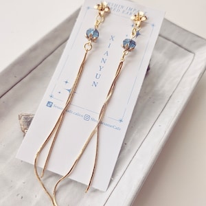 May include: A pair of gold chain earrings with blue crystal flower accents. The earrings are displayed on a white card with the text "XIANYUN" and "SUNSHINE IMPACT GOLD EARRINGS".