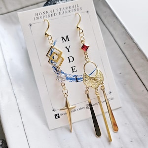 May include: Gold dangle earrings inspired by the Honkai Starrail video game. The earrings feature a red gemstone, a star, a crescent moon, and a geometric design. The earrings are displayed on a white card with the text "Honkai Starrail Inspired Earrings" and "MYDE".