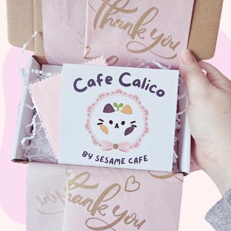 May include: A pink and white gift box with a white card that says "Cafe Calico by Sesame Cafe" and a cartoon cat with a pink bow. The box is filled with pink tissue paper and has a "thank you" message on the top.