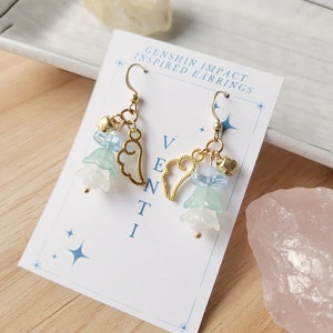 May include: Gold earrings inspired by the Genshin Impact character Venti. The earrings feature a heart charm, blue and white flower charms, and small bells.