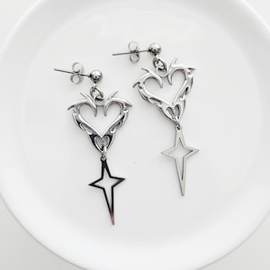 May include: A pair of silver-toned earrings. Each earring features a heart-shaped design with flame-like edges, a small ball, and a dangling star. The earrings are displayed on a white surface.