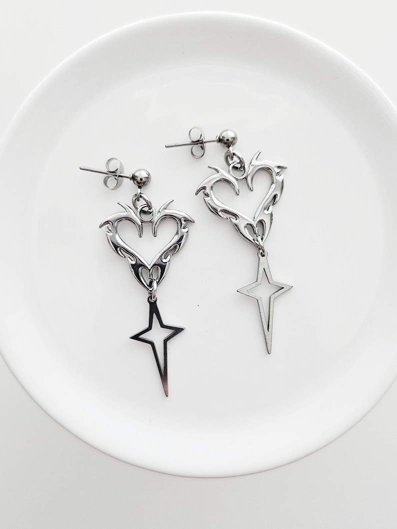 May include: A pair of silver-toned earrings. Each earring features a heart-shaped design with flame-like edges, a small ball, and a dangling star. The earrings are displayed on a white surface.