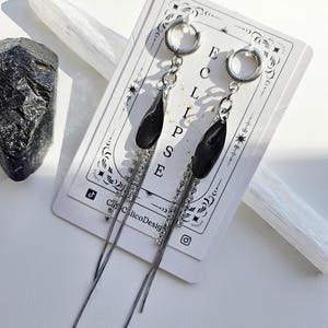 May include: Silver hoop earrings with black teardrop-shaped pendants and dangling silver chains. The earrings are displayed on a white card with the word "ECLIPSE" and the text "Cafe CalicoDesign".