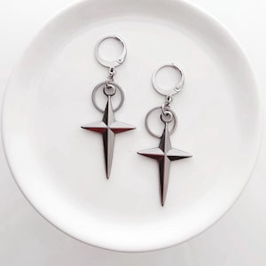 May include: A pair of silver-toned earrings featuring a four-pointed star design. Each earring has a circular hoop at the top, connected to a ring, and a star pendant. The earrings are displayed on a white, round plate.