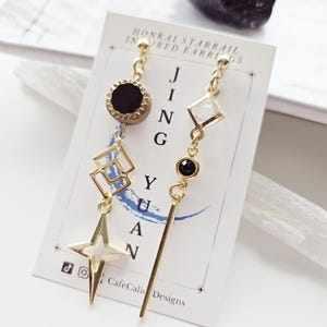 May include: Gold earrings inspired by the Honkai Starrail video game. The earrings feature a black stone, a geometric shape, and a star design. The earrings are on a white card with the text "Honkai Starrail Inspired Earrings" and "CafeCali Designs".