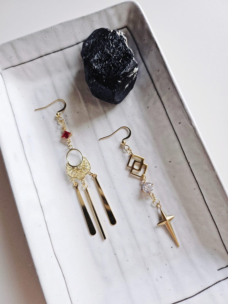 May include: A pair of gold earrings with dangling charms. One earring features a geometric design with a clear crystal and a star charm. The other earring has a circular design with a red stone and three dangling bars.