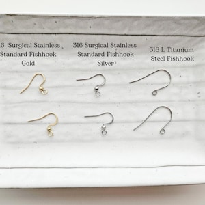 May include: A white rectangular tray displays six earring fishhooks. The top row features gold, silver, and steel hooks, each labeled with material and type. The bottom row mirrors the top, showcasing the same hook styles.