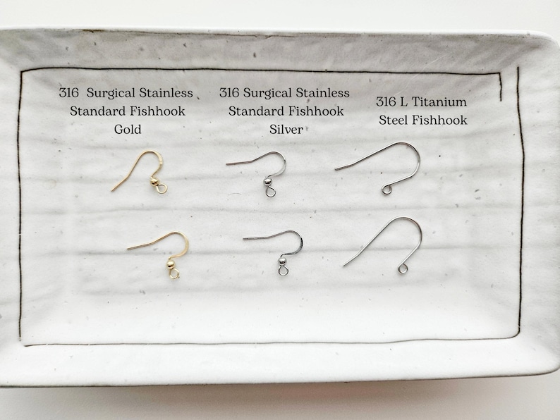 May include: A white rectangular tray displays six earring fishhooks. The top row features gold, silver, and steel hooks, each labeled with material and type. The bottom row mirrors the top, showcasing the same hook styles.