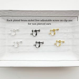 May include: A rectangular white ceramic tray displays six pairs of clip-on earrings in silver, black, and gold. The text on the tray reads: "Rack plated brass nickel free adjustable screw on clip ons for non pierced ears."