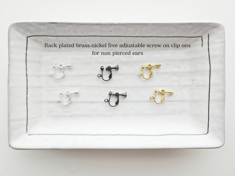 May include: A rectangular white ceramic tray displays six pairs of clip-on earrings in silver, black, and gold. The text on the tray reads: "Rack plated brass nickel free adjustable screw on clip ons for non pierced ears."
