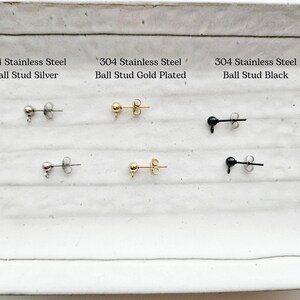 May include: A selection of ball stud earrings in silver, gold-plated, and black. Each pair is made of 304 stainless steel. The earrings are displayed on a white rectangular surface with text labels.