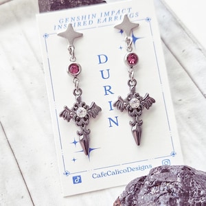 May include: A pair of Genshin Impact inspired earrings. Each earring features a star-shaped top, a pink gem, a cross-shaped design with a central clear gem, and a pointed bottom. The card behind the earrings reads "DURIN" and "CafeCalicoDesigns."