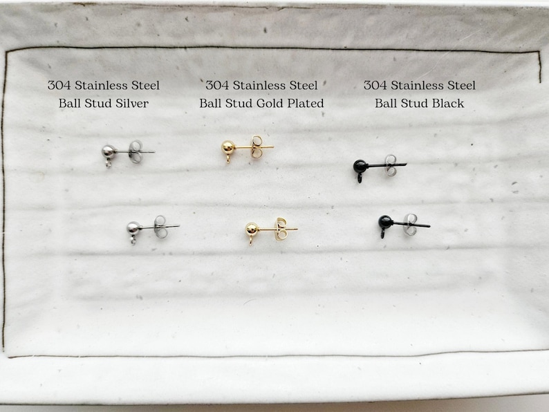 K&ouml;nnte beinhalten: Drei Paar 304 Edelstahl-Ball-Ohrstecker. Die Ohrringe werden auf einer wei&szlig;en Oberfl&auml;che gezeigt. Das erste Paar ist silber, das zweite Paar ist vergoldet und das dritte Paar ist schwarz.