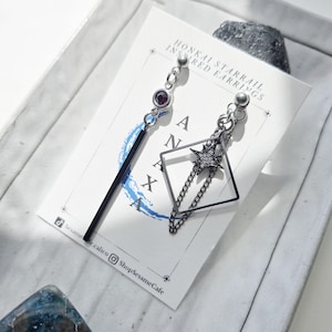 May include: Asymmetrical earrings. One earring has a black bar and a small purple gem. The other has a black square with a star and chain design. The earrings are on a white card with text. The card reads "Honkai Starrail Inspired Earrings".