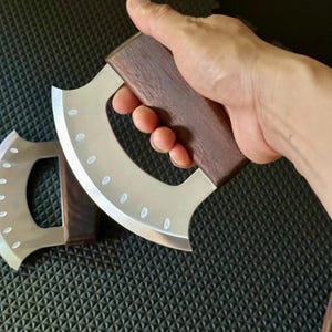 May include: A stainless steel mezzaluna knife with a curved blade and a dark wood handle. The blade has a series of small, evenly spaced holes. The knife is designed for chopping and mincing herbs and vegetables.