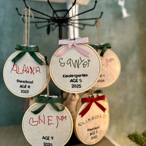 May include: Embroidered hoop ornaments with names and details like "Preschool AGE 4 2025" and "Kindergarten AGE 5 2025". Each ornament has a ribbon bow. The names are embroidered in red and black thread.