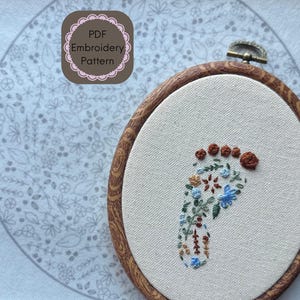 Peut inclure: Un patron de broderie PDF représentant un pied floral. La broderie est réalisée dans un cercle en bois, sur un tissu clair. Le pied est orné de fleurs bleues, marron et vertes. Le texte "PDF Embroidery Pattern" est en haut à gauche.