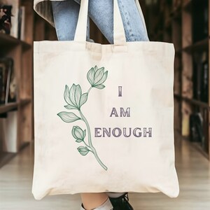 May include: Cream-colored tote bag with a green floral illustration and the text "I AM ENOUGH" in purple. The bag has a simple design and is held in front of a bookshelf.