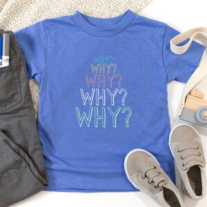 Op de afbeelding: Een blauw T-shirt voor peuters met het woord "WHY?" vijf keer herhaald in regenboogkleuren. Het shirt ligt op een wit oppervlak met een paar grijze sneakers, een houten speelgoedcamera en een grijze broek met een zak.