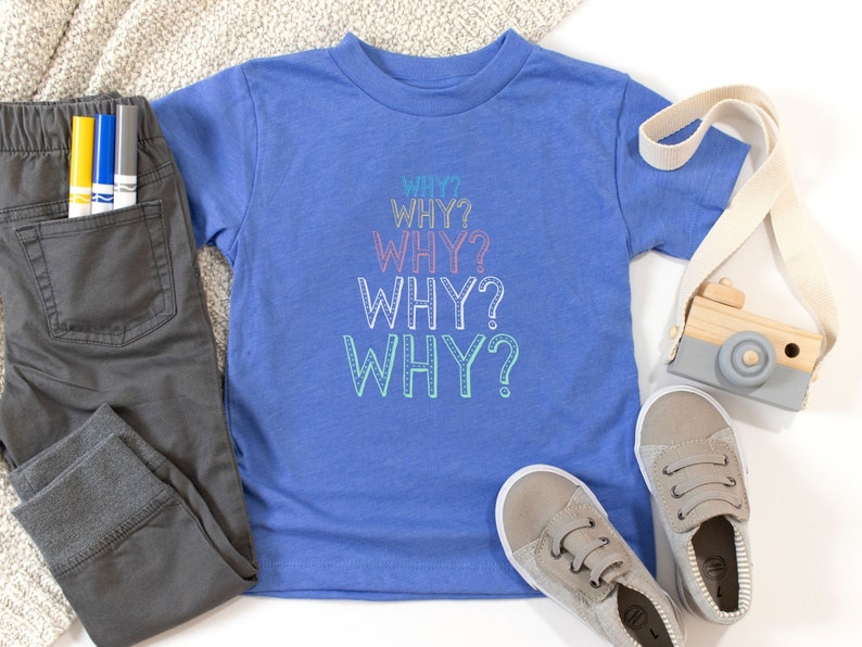 Op de afbeelding: Een blauw T-shirt voor peuters met het woord "WHY?" vijf keer herhaald in regenboogkleuren. Het shirt ligt op een wit oppervlak met een paar grijze sneakers, een houten speelgoedcamera en een grijze broek met een zak.