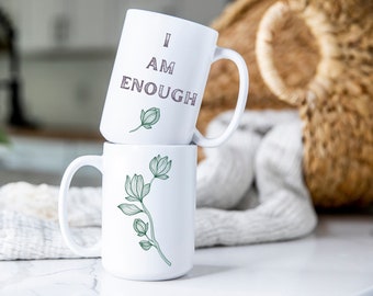 May include: Two white ceramic mugs stacked on top of each other. The top mug has the text "I AM ENOUGH" printed on it in a gray font. The bottom mug has a green floral design.