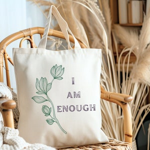 May include: Cream-colored tote bag with a floral design and the words "I AM ENOUGH" in purple. The bag is made of canvas and has two handles. The tote bag is sitting on a wicker chair.