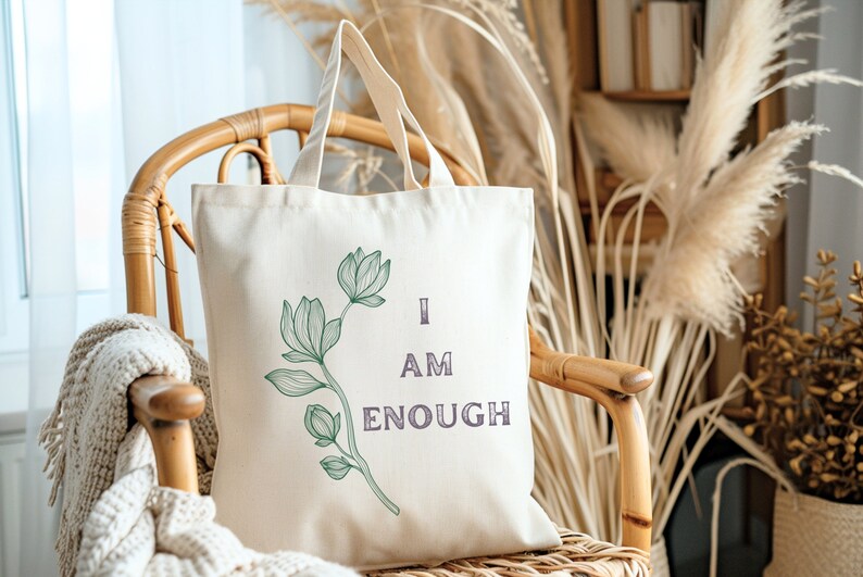 May include: Cream-colored tote bag with a floral design and the words "I AM ENOUGH" in purple. The bag is made of canvas and has two handles. The tote bag is sitting on a wicker chair.
