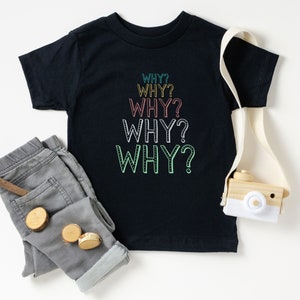 Op de afbeelding: Een zwart T-shirt met het woord "WHY?" vijf keer herhaald in een regenboog van kleuren. Het shirt ligt plat op een wit oppervlak met een grijze spijkerbroek en een houten speelgoedcamera.